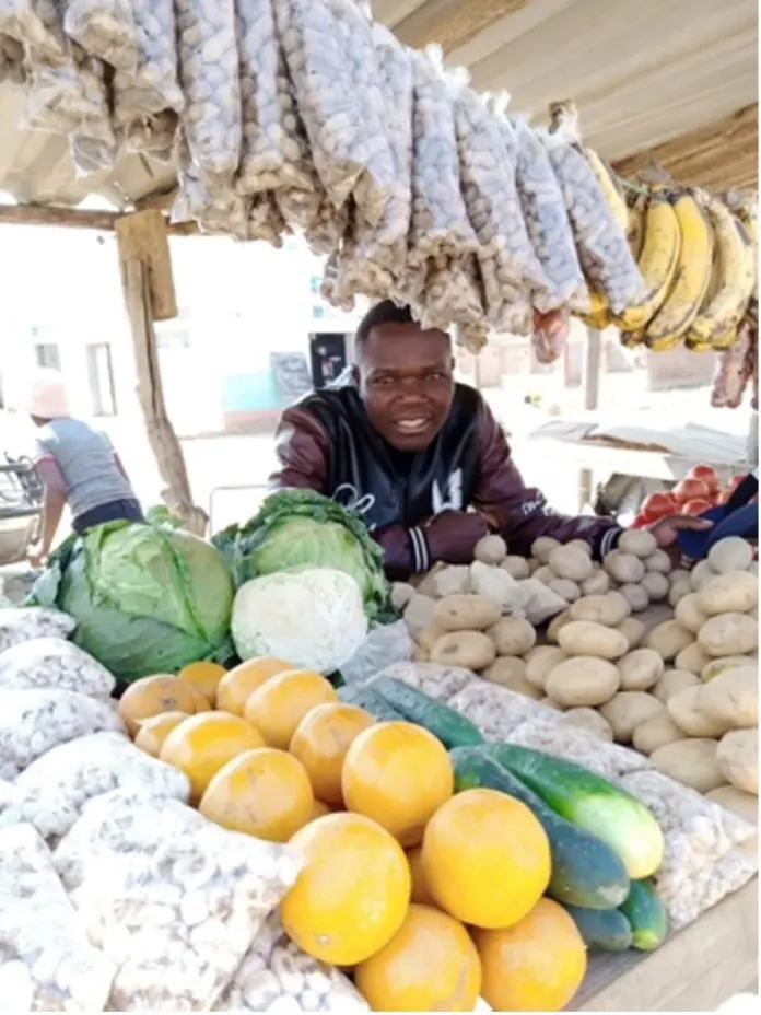 A young trader behind his market stall of cabbages, oranges, potatoes and cucumbers